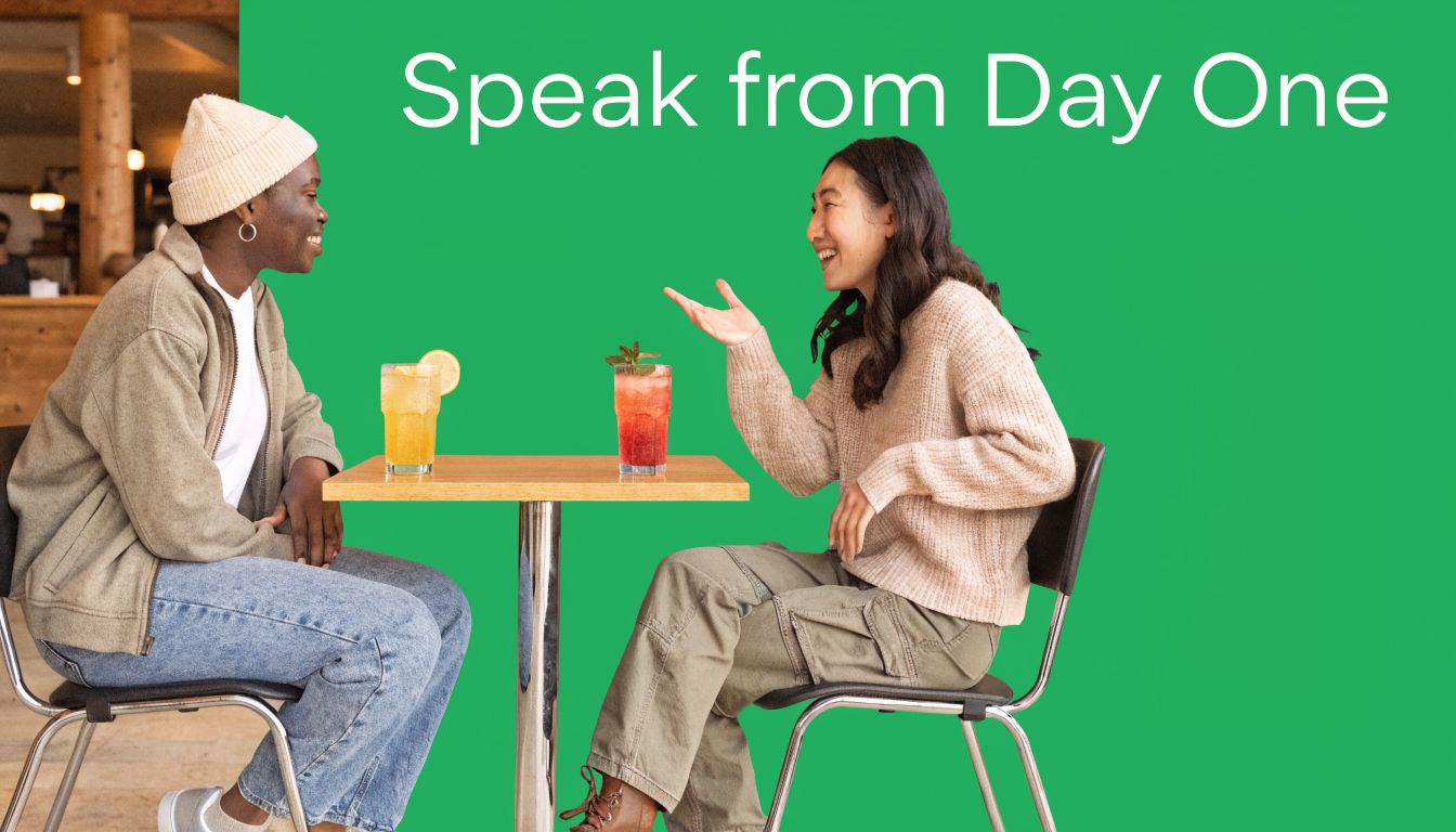 Two people talking and laughing while sitting at a cafe table with drinks on a green background.