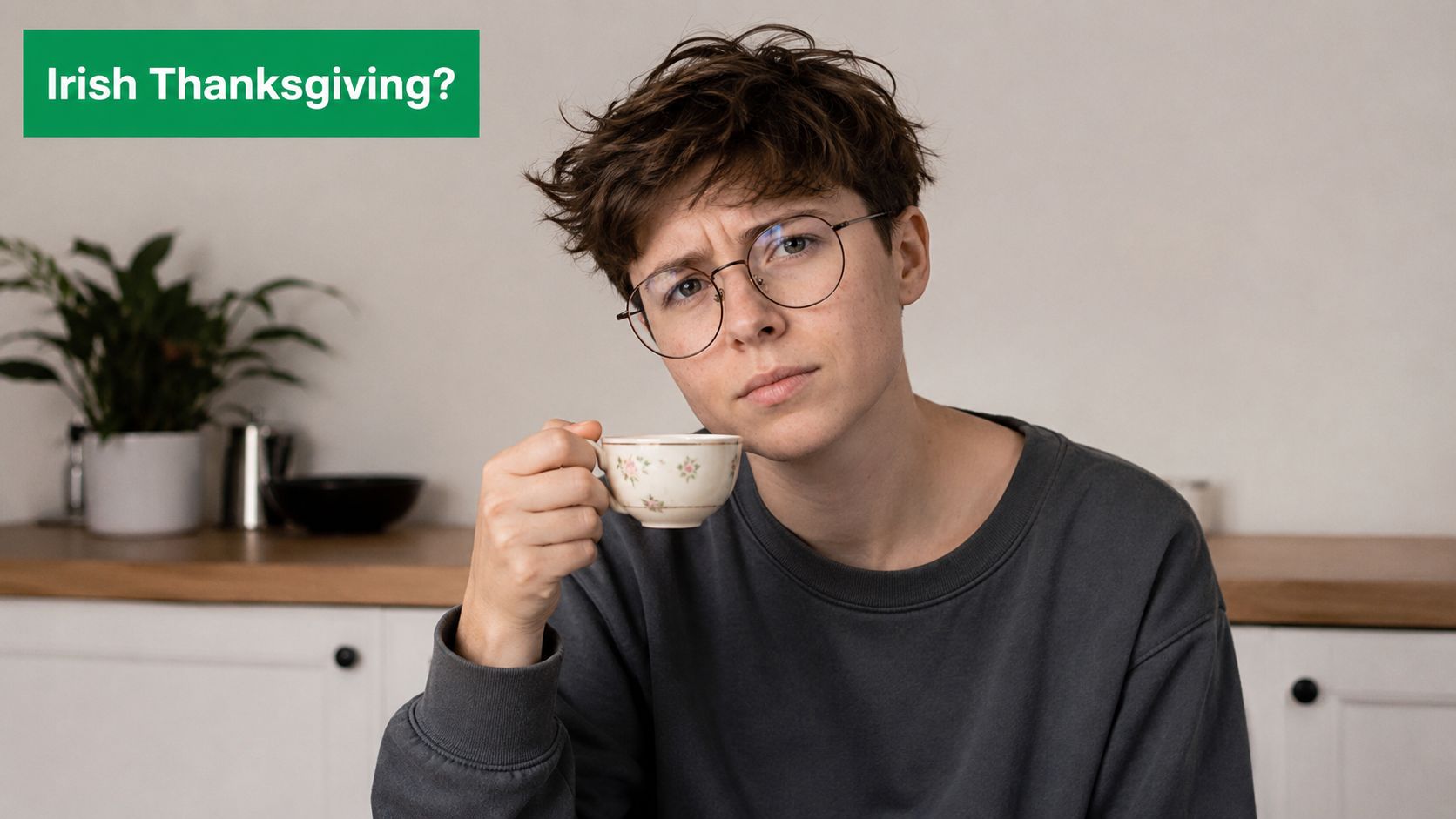 A person with short hair and glasses holding a small teacup and looking confused while sitting.