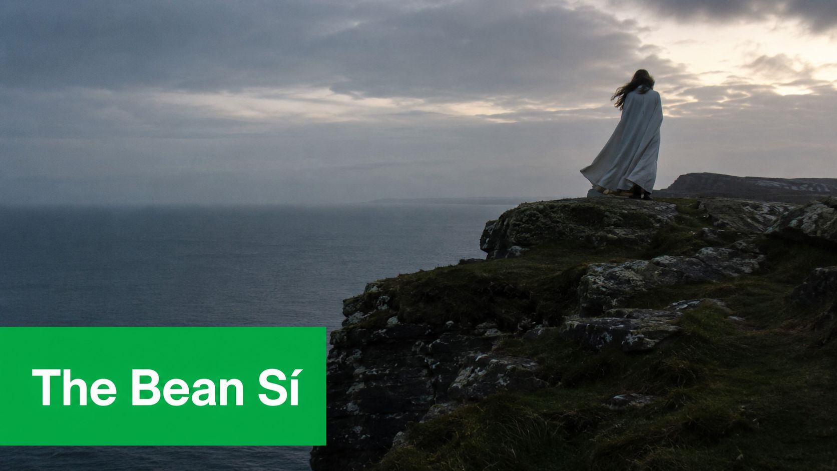 A mysterious figure in a long white cloak stands on a rugged cliff overlooking the ocean.